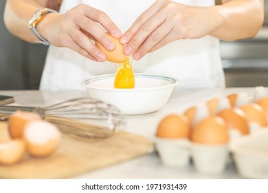 Young chef in white apron crack fresh egg yolk dropping in a bowl. Preparing raw ingredient for making healthy cuisine, depth of field. Hands holding eggshell with blurred cooking items. Homemade meal - Powered by Shutterstock