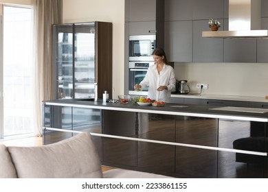 Young chef woman cooking salad in contemporary home kitchen interior, standing at island table, preparing organic food ingredients with modern appliance, furniture in background - Powered by Shutterstock