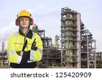Young chemical engineer posing in front of a biodiesel refinary plant, wearing a hard hat, fire retardant clothing with reflective stripes, looking proudly into the camera