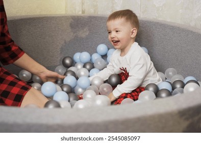 A young child with a big smile enjoys playing in a ball pit filled with various colored balls, while a caring adult sits nearby, creating a playful atmosphere of joy. - Powered by Shutterstock