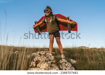 Young child with cardboard jetpack and wings standing on a rock, pretending to fly