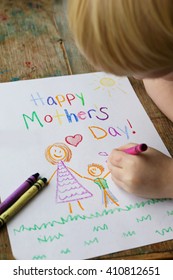 A young child is coloring a card with crayons for their mom that says Happy Mother's Day.