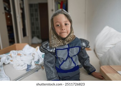 Young child dressed as a knight with toy sword, sitting on bed, imaginative play, wearing medieval costume, bedroom scene, childhood, creativity, playful, happy expression, knight armor - Powered by Shutterstock