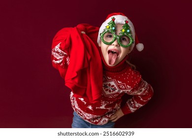 A young child dressed in a red Christmas sweater and playful green glasses with holiday decorations sticks out their tongue while holding a red cloth, exuding holiday joy. - Powered by Shutterstock