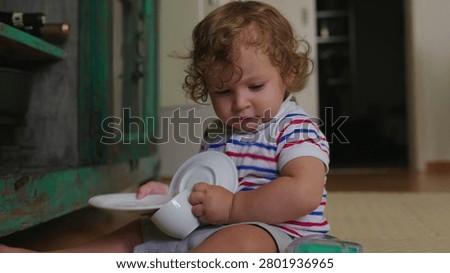 Young child examining breakable dishware while seated on floor unsafe interaction moment highlighting curiosity and potential danger with fragile objects indoors