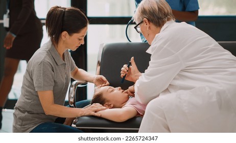 Young child fainting in hospital waiting room, elderly doctor doing emergency consultation with flashlight. Worried mother supporting little kid being sick and unconscious. Handheld shot. - Powered by Shutterstock