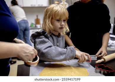 A young child, focused and determined, rolls out dough with a rolling pin while using a cookie cutter. Two adults assist nearby in a warm, inviting kitchen filled with baking tools. Christmas craft - Powered by Shutterstock