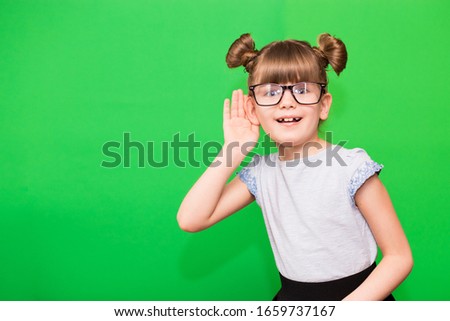 A young child girl journalist in glasses with funny pigtails listening to something holding his hand to ear isolated on green background.