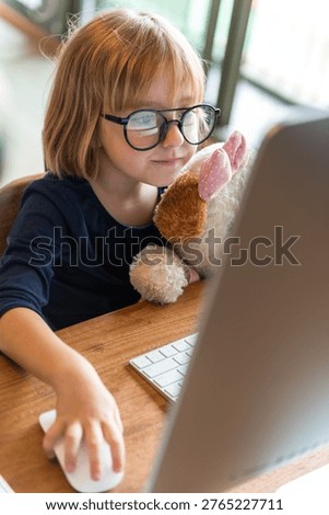 Young child with glasses using a computer, holding a stuffed animal. The child is focused on the screen, creating a cozy and studious atmosphere.