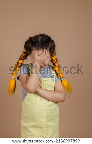 Young child having yellow kanekalon pigtails showing feelings of shame, touching forehead with hand in yellow jumpsuit and gray t-shirt on beige background.
