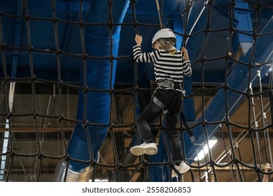 A young child in a helmet scales a climbing net at an indoor ropes course, demonstrating skills and excitement in a safe, fun environment - Powered by Shutterstock