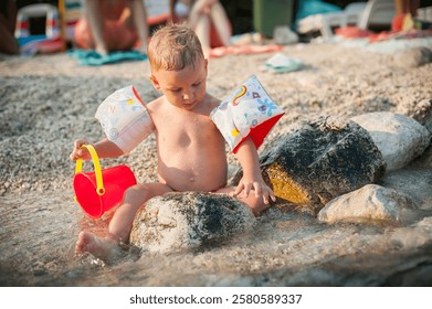 A young child with inflatable arm floaties sits on rocks by the water’s edge, splashing and playing with a small red bucket. The warm sun creates a joyful atmosphere.