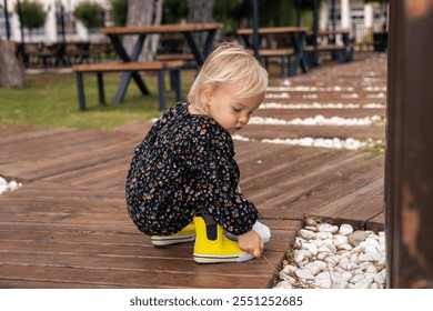 A young child kneels on a wooden path in a park after rain, examining white pebbles while wearing a floral outfit and bright yellow shoes. The scene captures a moment of curiosity and exploration. - Powered by Shutterstock