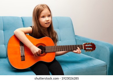 Young Child, Little School Age Girl Playing Classic Acoustic Guitar, Practicing Musical Instrument Sitting On The Sofa Simple Music Education At Home, Child's Hobbies And Talents, Portrait, Interior. 