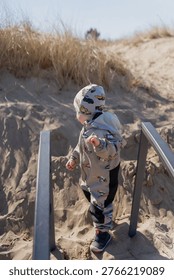 Young child in patterned hat standing on beach dune staircase with metal rails, seaside access, outdoor activity and family leisure theme