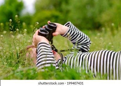 Young Child Using A Pair Of Binoculars To Look Up Into The Sky As He Lies On Back In A Grassy Rural Meadow Enjoying A Day In Nature