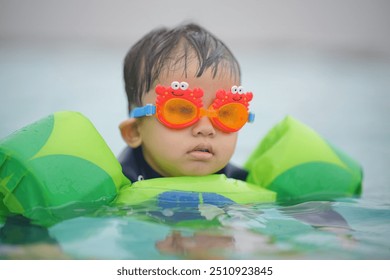 Young child wearing flotation devices and colorful goggles in a pool, learning to swim and enjoying water activities. - Powered by Shutterstock