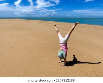 Young child wearing a sun hat doing a cartwheel on an empty long sandy beach creating a dark shadow on the ground - Powered by Shutterstock