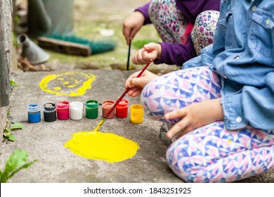 Young Children Painting On Concrete, Two Anonymous Girls Engaging In Art Therapy Activities. Small Kids Spreading Colorful Paint On The Ground Using A Brush, Outdoors Scene, Closeup. Kids And Art