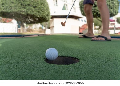 Young Child's Feet On A Minature Or Mini Golf Hole Close Up With Ball Falling To The Hole On Green Artificial Turf Outdoors.