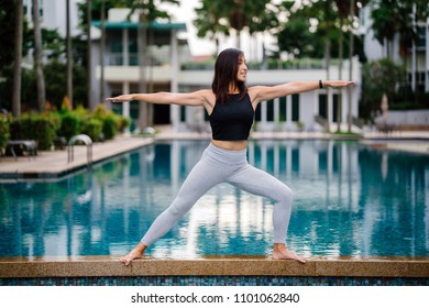 A young Chinese Asian girl is in a yoga pose (warrior) by the poolside. She is a Singaporean millennial teen and is smiling as she stretches and goes into the pose. 