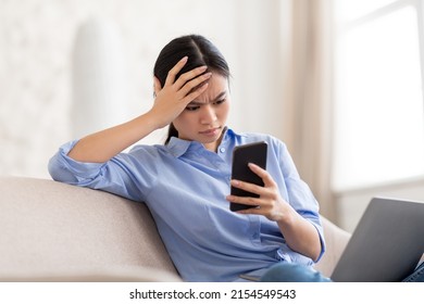 Young Chinese Businesswoman Sitting On Couch, Using Laptop And Smartphone At Home, Angry Asian Lady Looking At Cell Phone Screen While Working Online, Reading Bad News, Copy Space