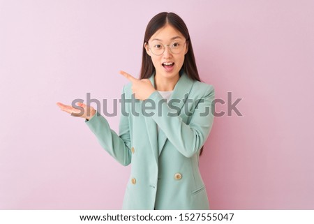 Young chinese businesswoman wearing jacket and glasses over isolated pink background amazed and smiling to the camera while presenting with hand and pointing with finger.
