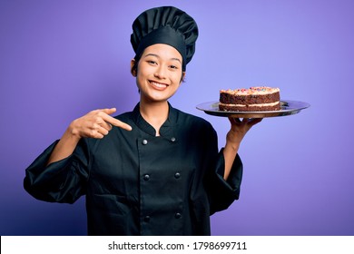 Young Chinese Chef Woman Wearing Cooker Uniform And Hat Holding Tray With Cake With Surprise Face Pointing Finger To Himself