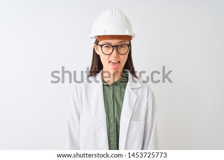 Young chinese engineer woman wearing coat helmet glasses over isolated white background sticking tongue out happy with funny expression. Emotion concept.