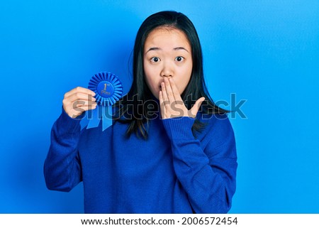 Young chinese girl holding first place badge covering mouth with hand, shocked and afraid for mistake. surprised expression 