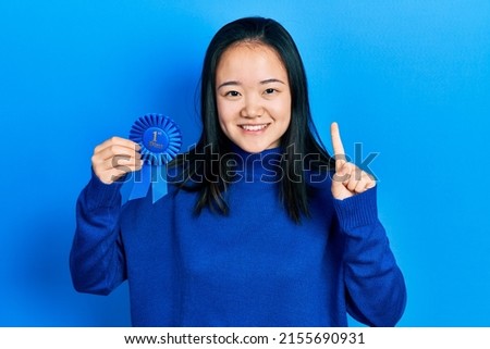 Young chinese girl holding first place badge smiling with an idea or question pointing finger with happy face, number one 