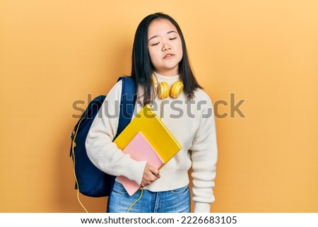 Young chinese girl holding student backpack and books looking sleepy and tired, exhausted for fatigue and hangover, lazy eyes in the morning. 