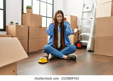 Young Chinese Girl Sitting On The Floor At New Home Covering Ears With Fingers With Annoyed Expression For The Noise Of Loud Music. Deaf Concept. 