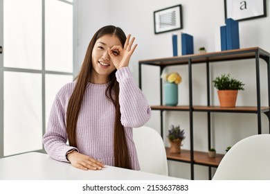Young chinese girl wearing casual clothes sitting on the table at home smiling happy doing ok sign with hand on eye looking through fingers 