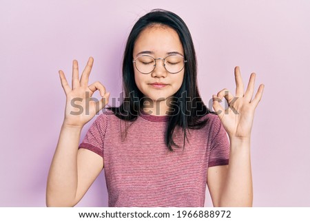 Young chinese girl wearing casual clothes and glasses relax and smiling with eyes closed doing meditation gesture with fingers. yoga concept. 