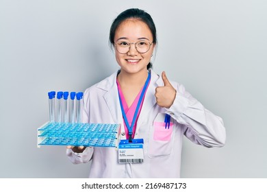 Young Chinese Girl Wearing Scientist Uniform Holding Test Tube Smiling Happy And Positive, Thumb Up Doing Excellent And Approval Sign 