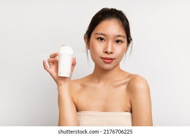 Young Chinese Lady Wrapped In Towel Showing Deodorant, Posing After Shower On White Studio Background. Body Care, Armpits, Underarms Hygiene Concept