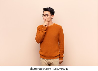 young chinese man asking for silence   quiet  gesturing and finger in front mouth  saying shh keeping secret against flat color wall