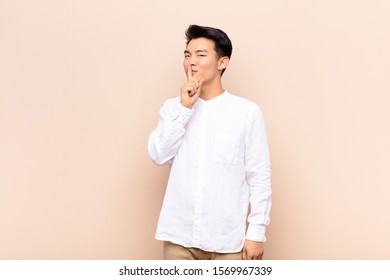 young chinese man asking for silence   quiet  gesturing and finger in front mouth  saying shh keeping secret against flat color wall