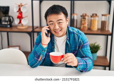 Young Chinese Man Drinking Coffee And Talking On The Smartphone At Home