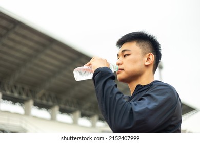 Young Chinese Man Drinking Water After Workout Exercising In The Stadium. Young Asian Man Drinking Water After Jogging Running Outdoor. Healthy And Active Lifestyle Concept.