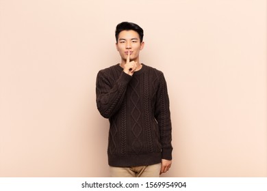 young chinese man looking serious   cross and finger pressed to lips demanding silence quiet  keeping secret against flat color wall