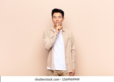 young chinese man looking serious   cross and finger pressed to lips demanding silence quiet  keeping secret against flat color wall