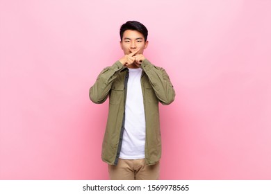 young chinese man looking serious   displeased and both fingers crossed up front in rejection  asking for silence against flat color wall