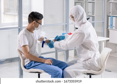 Young Chinese Man In Mask Sitting In Lab While Doctor In Protective Coveralls Taking His Blood In Syringe For Analysis