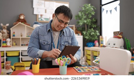 Young chinese man preschool teacher using touchpad sitting on table at kindergarten - Shutterstock ID 2261369819