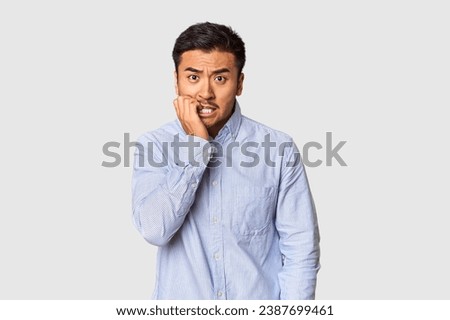 Young Chinese man in studio background biting fingernails, nervous and very anxious.