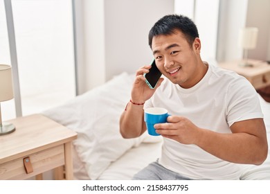 Young Chinese Man Talking On The Smartphone Drinking Coffee Sitting On Bed At Bedroom