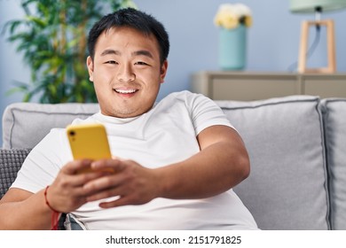 Young Chinese Man Using Smartphone Sitting On Sofa At Home