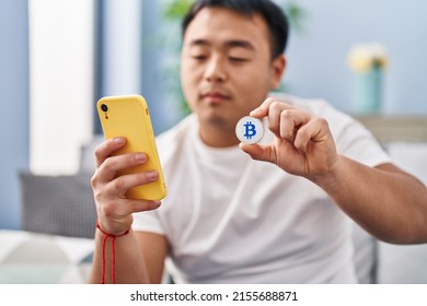 Young Chinese Man Using Smartphone Holding Bitcoin At Home